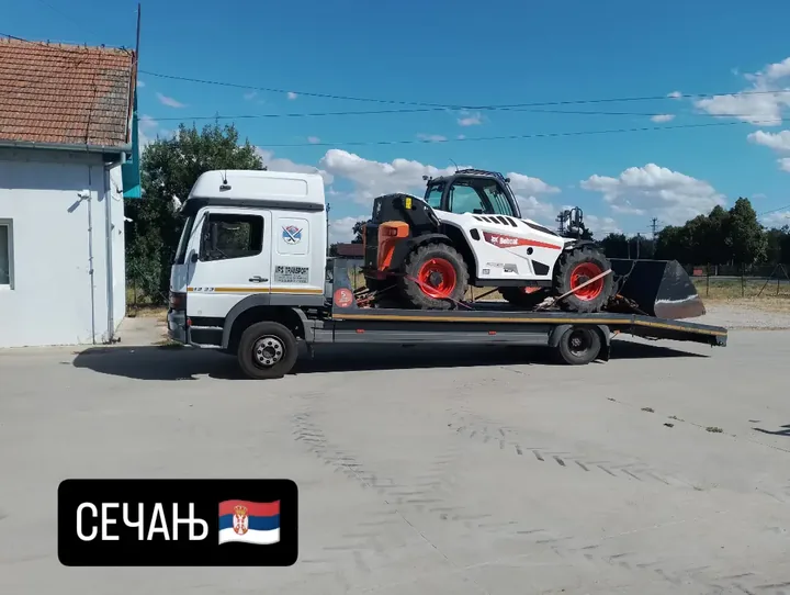 Beli kompaktni utovarivač na transportnoj platformi kamiona.